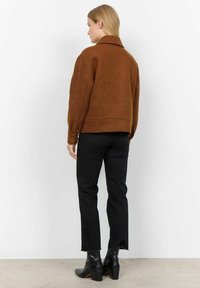 Veste en tissu marron, douce, avec un col, un design court et des poignets boutonnés, associée à un pantalon noir slim et des bottines.