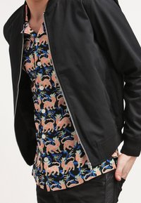 Chaqueta bomber negra sobre una camisa de botones colorida con un patrón de tigre sobre un fondo negro, con acentos en azul y verde.