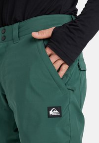 Grüne Outdoor-Hosen mit einem eleganten Design, hergestellt aus wasserdichtem Material. Sie verfügen über eine Fronttasche und ein Quiksilver-Logo-Patch auf dem Oberschenkel.