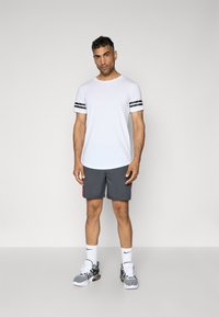 Hvid crew-neck t-shirt med sorte striber på ærmerne, parret med grå shorts med lyserøde detaljer. Modellen har grå og sorte løbesko på.