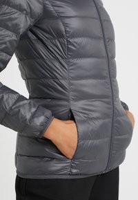 Chaqueta acolchada gris con costuras horizontales, cierre frontal con cremallera y bolsillos laterales. Presenta una textura suave y reflectante y mangas ajustadas.