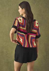 Bunt gehäkeltes Oberteil mit kurzen Ärmeln, das geometrische Muster in Orange, Pink, Gelb und Blau zeigt, kombiniert mit schwarzen Shorts.