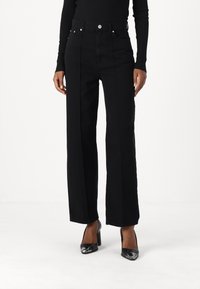 Hooggetailleerde zwarte denim wijde broek met voornaad, vijf zakken en zilverkleurige hardware, gecombineerd met zwarte hoge hakken.