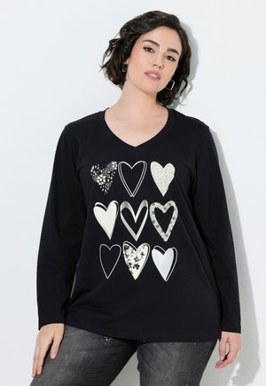 Mujer con el pelo corto y oscuro que lleva una camiseta negra de manga larga con nueve diferentes diseños de corazones blancos en la parte delantera, de pie frente a una pared blanca.