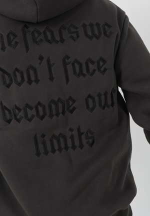 Sudadera negra con capucha que presenta bordado en relieve en negro con el texto "Los miedos que no enfrentamos se convierten en nuestros límites" en la parte posterior. Textura de tela suave.