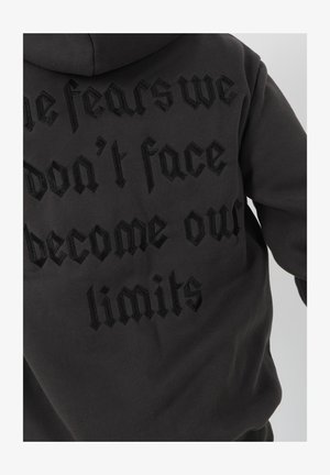 Sudadera negra con capucha que presenta bordado en relieve en negro con el texto "Los miedos que no enfrentamos se convierten en nuestros límites" en la parte posterior. Textura de tela suave.