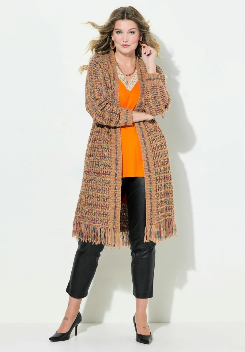 Cardigan en tricot beige avec des éclats multicolores, manches longues, ouvert à l'avant et ourlet à franges, associé à un haut orange et un pantalon noir.