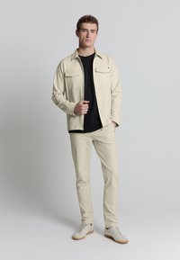 Beige lichtgewicht jack met twee voorzakken, gedragen over een zwart t-shirt, gecombineerd met beige broek en witte sneakers op een neutrale achtergrond.