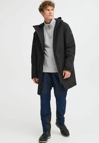 Črna vodoodporna jakna s kapuco, z sprednjimi žepi in zadrgo. Nošena nad sivo majico z dolgimi rokavi in temno modrimi cargo hlačami.