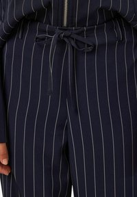 Marineblå bukser i stoff med hvite vertikale striper. Har knytebelte i midjen og myk tekstur. Lynlås på toppen.