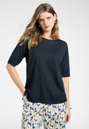 Kvinde med langt blondt hår iført en løs mørkeblå skjorte og hvide bukser med blå og gule abstrakte fjer-mønstre.