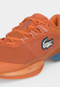 Πορτοκαλί αθλητικό παπούτσι Lacoste με μπλε λογότυπο αλιγάτορα, επάνω μέρος από διχτυωτό ύφασμα, επενδεδυμένο κολάρο και ορατή λεπτομέρεια σόλας με απορρόφηση κραδασμών LSRFOAM.
