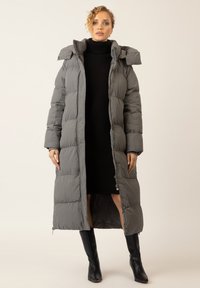 Langer grauer Daunenmantel mit Kapuze, mit gestepptem Design, einem Reißverschluss vorne und seitlichen Taschen, getragen über einem schwarzen Kleid und knielangen Stiefeln.