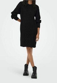 Robe noire en maille présentant un motif texturé, un col rond et des manches longues. Portée avec des bottines noires épaisses.