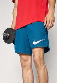 Person wearing blue Nike shorts and red shirt, holding a black dumbbell in their left hand against a plain background.