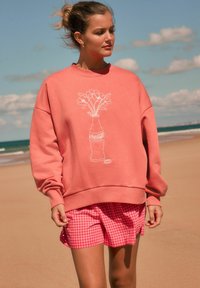 Korallefarbener Pullover mit weißem grafischem Blumenvasenmotiv, kombiniert mit rosa Karoshorts. Aus weichem Baumwollmaterial, lockerer Schnitt und gerippte Bündchen.