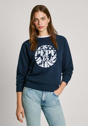 Felpa blu navy con testo grafico bianco, polsini e orlo a costine, colletto rotondo, indossata con jeans azzurri strappati.