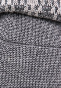 Nahaufnahme eines gefalteten grauen Strickstoffs mit einem strukturierten Muster und einem überlappenden Abschnitt, der ein cremfarbenes und graues gesticktes Design zeigt.