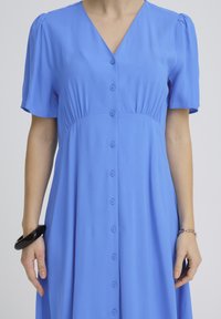 Woman wearing a blue button-down dress with short sleeves, a black bracelet on left wrist, and a silver chain bracelet on right wrist.