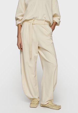 Osoba ubrana w luźne kremowe spodnie z dopasowanym paskiem, luźną kremową bluzkę z podwiniętymi rękawami oraz kremowe wsuwane buty.