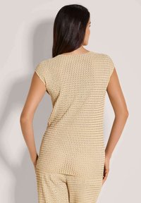 Vrouw met lang donker haar die een beige textuur korte mouwen top en bijpassende broek draagt, van achteren gezien tegen een effen achtergrond.