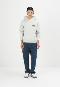 Lys grå hettegenser med en teksturert finish, frontlomme og logo-accent. Kombinert med mørkeblå jeans og hvite sneakers med grå detaljer.