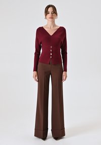 Maglione a maglia bordeaux con scollo a V e tre bottoni argentati, abbinato a pantaloni a vita alta in marrone con gamba ampia e orlo piegato.