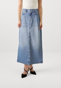 Jupe en denim bleu clair, mi-longue, avec des détails de couture visibles. Dotée d'une fermeture à bouton à l'avant et de passants de ceinture, portée avec des talons noirs.