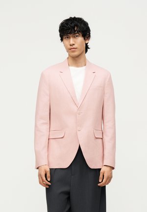 Joven con pelo negro y rizado que lleva una chaqueta rosa claro sobre una camisa blanca y pantalones gris oscuro sobre un fondo blanco liso.