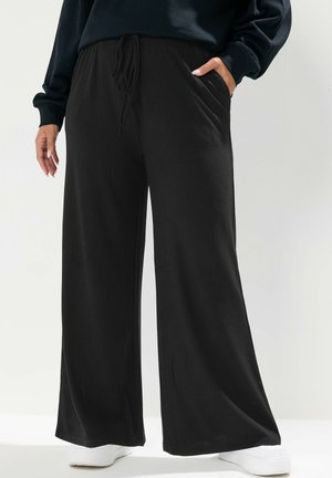 Persoon draagt losse zwarte geribbelde wijde broek met trekkoord in de taille en witte sneakers, één hand in de zak en één ontspannen langs het lichaam.
