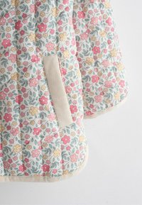 Gequilte stof met een roze, gele en rode bloemenprint en een beige geribbelde zakdetail op een witte achtergrond.