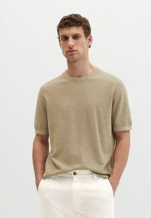 Mężczyzna w beżowym, krótkim swetrze z dzianiny i białych spodniach, stojący z rękami w kieszeniach na jednolitym białym tle.