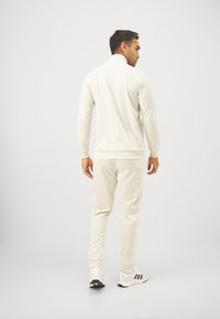 Biely športový komplet vyrobený z mäkkého materiálu. Obsahuje golier na zips, elastické manžety a postranné pruhy na nohaviciach.