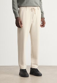 Pantalons beige à taille élastique avec détail de cordon, coupe ajustée, texture lisse et poches latérales, associés à des baskets noires.
