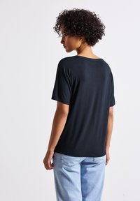 T-shirt nera a maniche corte con vestibilità rilassata, caratterizzata da un colletto rotondo e una texture liscia, abbinata a jeans in denim blu chiaro.