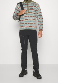 Pullover en polaire à motif multicolore avec un zip demi-long et des accents gris, associé à un pantalon noir fuselé avec une poche latérale. Chaussures noires.