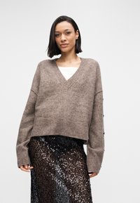 Καφέ πουλόβερ με V λαιμόκοψη από απαλό υλικό, με ριμπ τελείωμα και μακριά μανίκια με διακοσμητικά κουμπιά, σε συνδυασμό με μαύρη φούστα με παγιέτες.