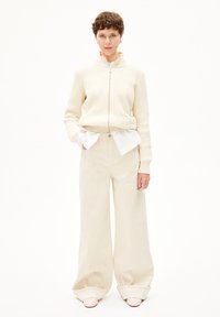 Crème ribbelige zip-up jacket over een witte overhemd, gecombineerd met beige wijde broek en lichtroze schoenen.