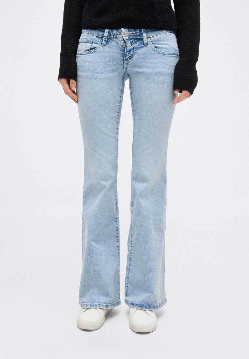 Άτομο που φοράει ανοιχτό μπλε τζιν με καμπάνα, λευκά αθλητικά παπούτσια και μαύρο μακρυμάνικο πουλόβερ, στέκεται σε μονόχρωμο φόντο.