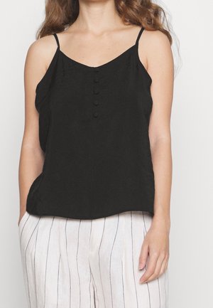 Frau trägt ein schwarzes ärmelloses Oberteil mit Knöpfen vorne und weiße Hosen mit dünnen vertikalen Streifen, steht mit den Händen in den Taschen.