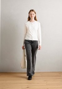 Bijela cardigan s gumbima, uparena s tamno sivim širokim hlačama i crnim cipelama. Osoba nosi torbu svijetle boje. Jednostavna pozadina.