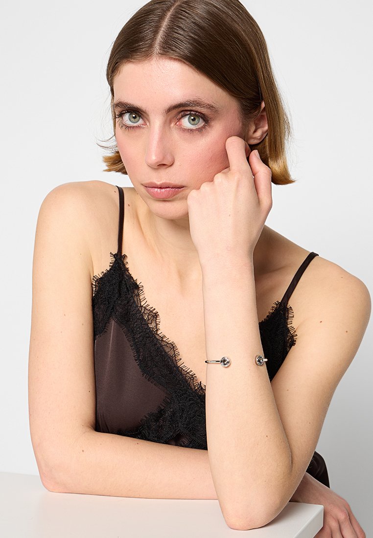 Pulsera de plata con dos piedras circulares, llevaba en una muñeca. El modelo viste una blusa de satén marrón con encaje negro. Fondo brillante.