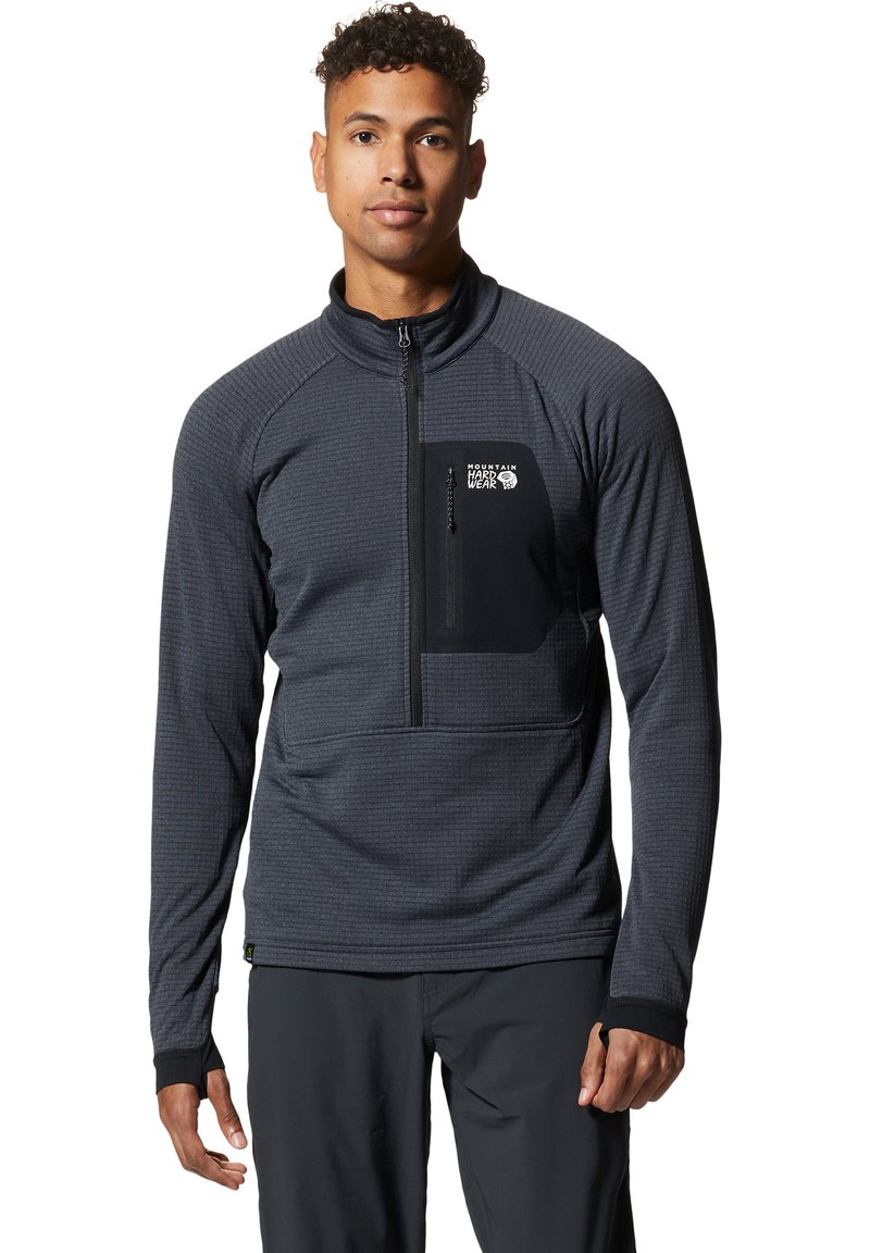 Młody mężczyzna w ciemnoszarym swetrze z połową zamka, z czarną kieszenią na zamek na klatce piersiowej oraz logiem "Mountain Hardwear".