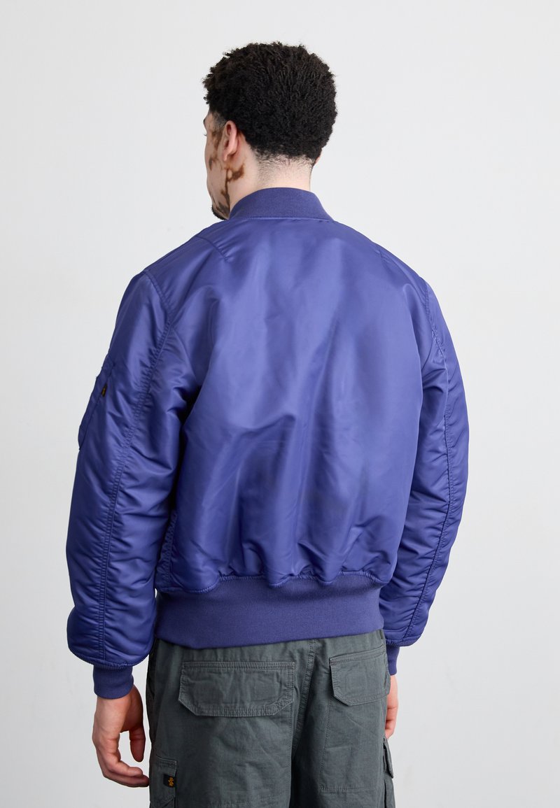 Giacca bomber viola con una texture liscia e lucida, polsini e vita a costine. Presenta un design aderente e una silhouette leggermente croppata.