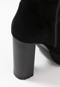 Bottine en daim noir avec un talon haut chunky et une fermeture éclair visible sur le côté, photographiée sur un fond blanc.