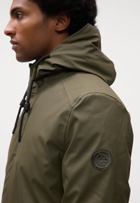 Olivengrøn vandtæt jakke med høj krave, justerbar hætte og et sort logo-patch på venstre skulder. Har en glat tekstur.