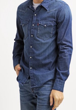 Homme portant une chemise en denim bleu foncé ajustée avec boutons et poches à rabats, et un jean bleu, debout avec une main dans la poche.