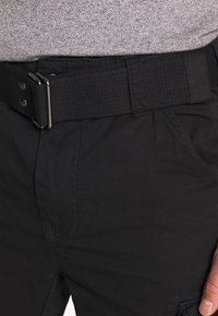 Zwarte cargobroek van een textuurstof, met een brede zwarte geweven riem met een metalen sluiting en zichtbare stiksels.