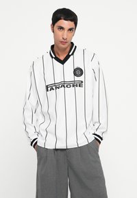 Jersey blanc à manches longues avec des rayures verticales noires, col en V ajusté et poignets côtelés. Présente un logo et le nom de la marque sur le devant.