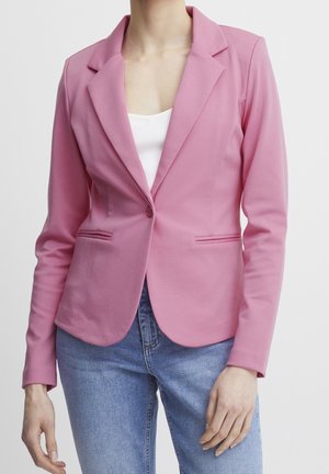 Frau trägt einen taillierten pinkfarbenen Blazer mit Revers über einem weißen Oberteil und blauen Jeans, stehend vor einem einfarbigen Hintergrund.